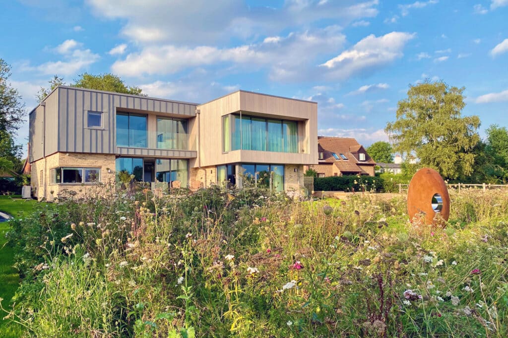 Modern house with large windows situated behind a wildflower garden. A circular sculpture stands in the foreground. Cloudy sky and neighboring houses in the background.