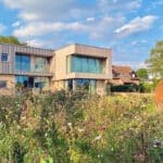 Thumbnail of http://Modern%20house%20with%20large%20windows%20situated%20behind%20a%20wildflower%20garden.%20A%20circular%20sculpture%20stands%20in%20the%20foreground.%20Cloudy%20sky%20and%20neighboring%20houses%20in%20the%20background.
