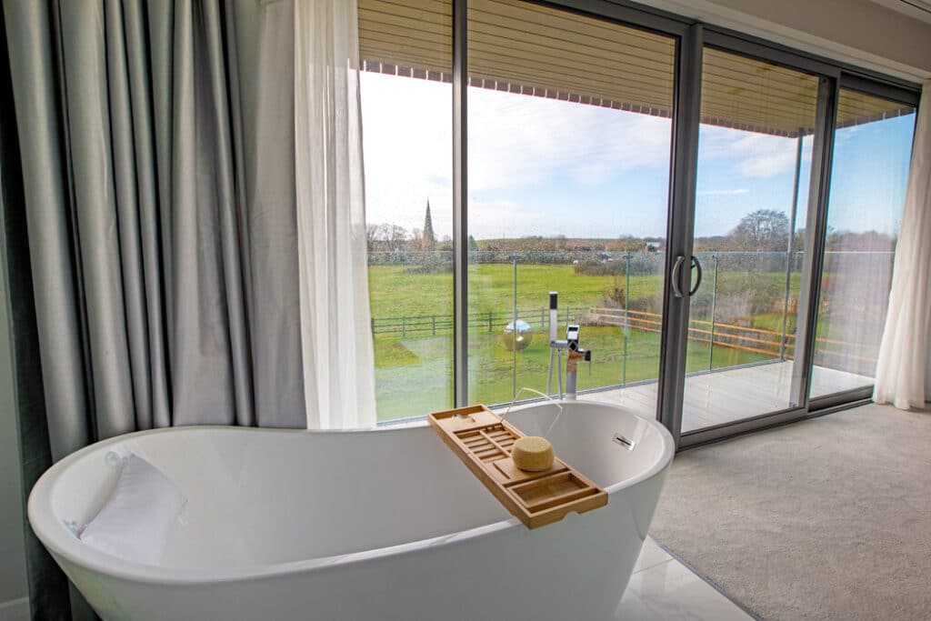 Freestanding bathtub near large glass doors with view of a green landscape and distant spire, inside a room with carpeted floor and gray curtains.