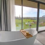 Thumbnail of http://Freestanding%20bathtub%20near%20large%20glass%20doors%20with%20view%20of%20a%20green%20landscape%20and%20distant%20spire,%20inside%20a%20room%20with%20carpeted%20floor%20and%20gray%20curtains.