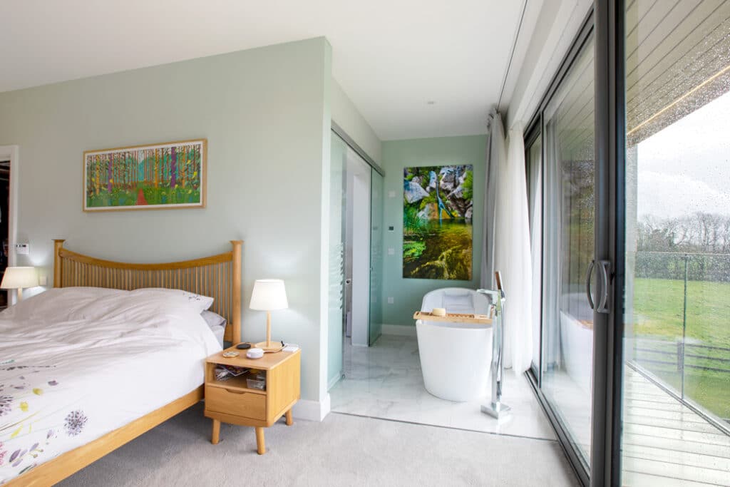 Bedroom with a double bed, nightstand, and lamp. Open bathroom with tub and artwork. Large sliding glass doors lead to a balcony with a garden view. Walls are light green.