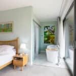 Thumbnail of http://Bedroom%20with%20a%20double%20bed,%20nightstand,%20and%20lamp.%20Open%20bathroom%20with%20tub%20and%20artwork.%20Large%20sliding%20glass%20doors%20lead%20to%20a%20balcony%20with%20a%20garden%20view.%20Walls%20are%20light%20green.