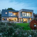 Thumbnail of http://Modern%20two-story%20house%20with%20large%20windows,%20surrounded%20by%20greenery.%20There%20is%20a%20circular%20metal%20sculpture%20on%20the%20lawn,%20and%20neighboring%20traditional-style%20house%20visible%20in%20the%20background.