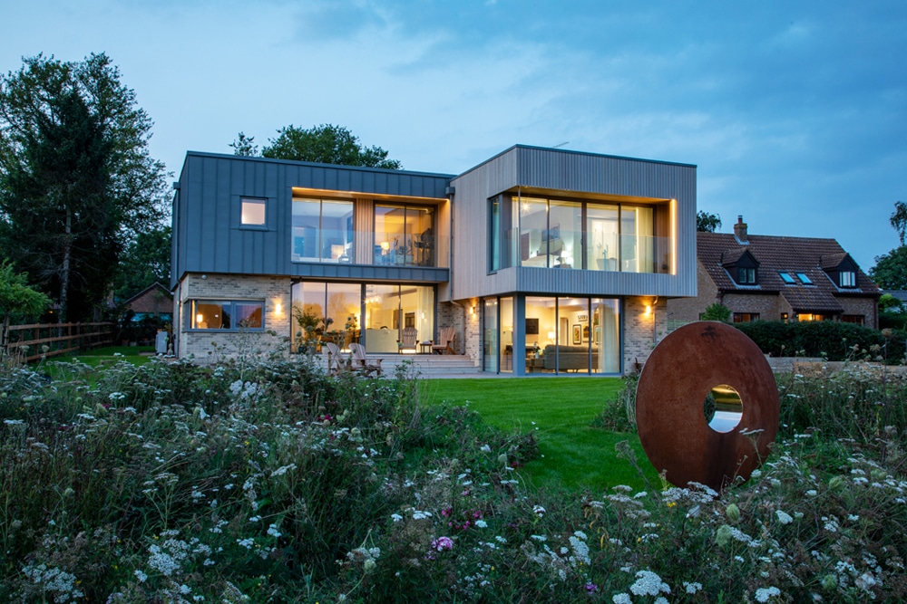 Modern two-story house with large windows, surrounded by greenery. There is a circular metal sculpture on the lawn, and neighboring traditional-style house visible in the background.