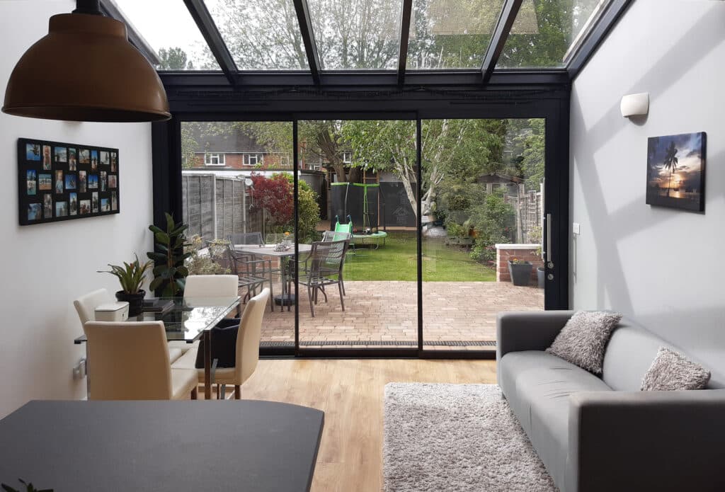 Modern living room with a grey sofa, dining area, and large glass doors opening to a backyard with outdoor furniture. White walls have photos and a scenic painting.