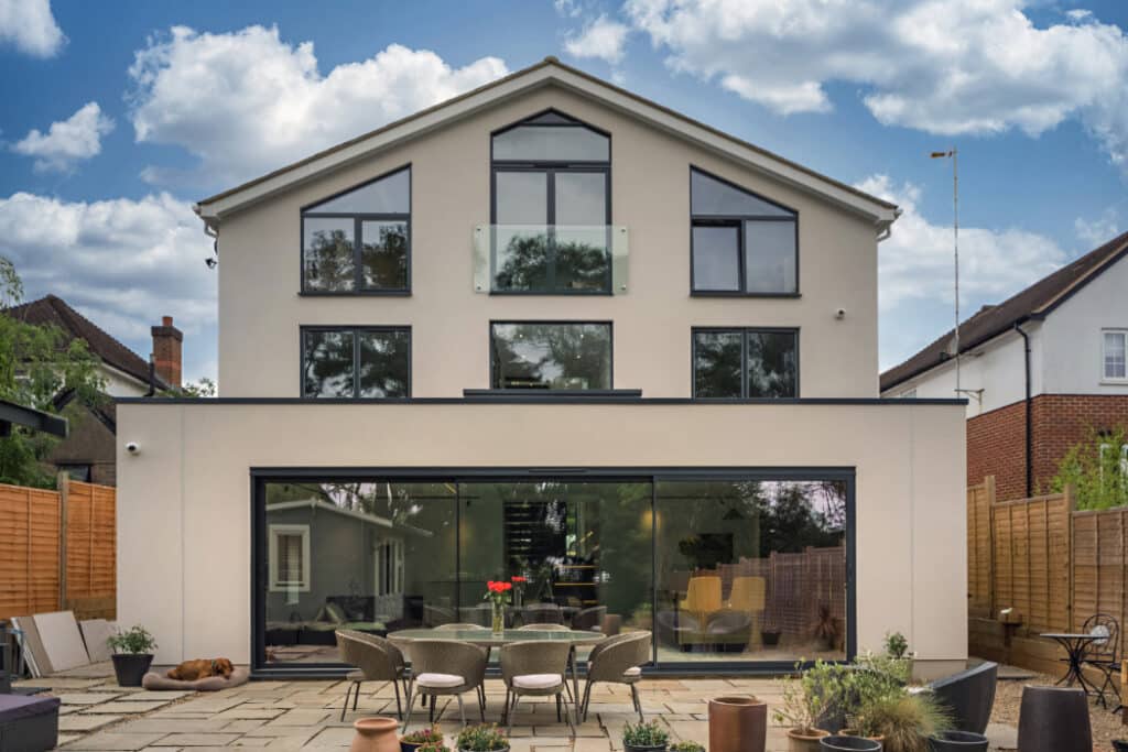 Modern two-story house with large glass windows, outdoor patio set with chairs and table, potted plants, and cloudy sky in the background.
