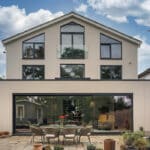 Thumbnail of http://Modern%20two-story%20house%20with%20large%20glass%20windows,%20outdoor%20patio%20set%20with%20chairs%20and%20table,%20potted%20plants,%20and%20cloudy%20sky%20in%20the%20background.