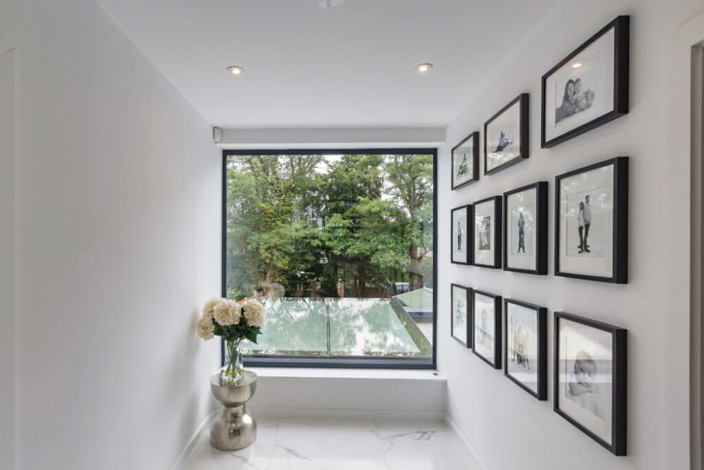 A hallway with white walls and a large window shows framed photos and a silver vase of flowers. There is a view of trees outside.