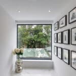 Thumbnail of http://A%20hallway%20with%20white%20walls%20and%20a%20large%20window%20shows%20framed%20photos%20and%20a%20silver%20vase%20of%20flowers.%20There%20is%20a%20view%20of%20trees%20outside.