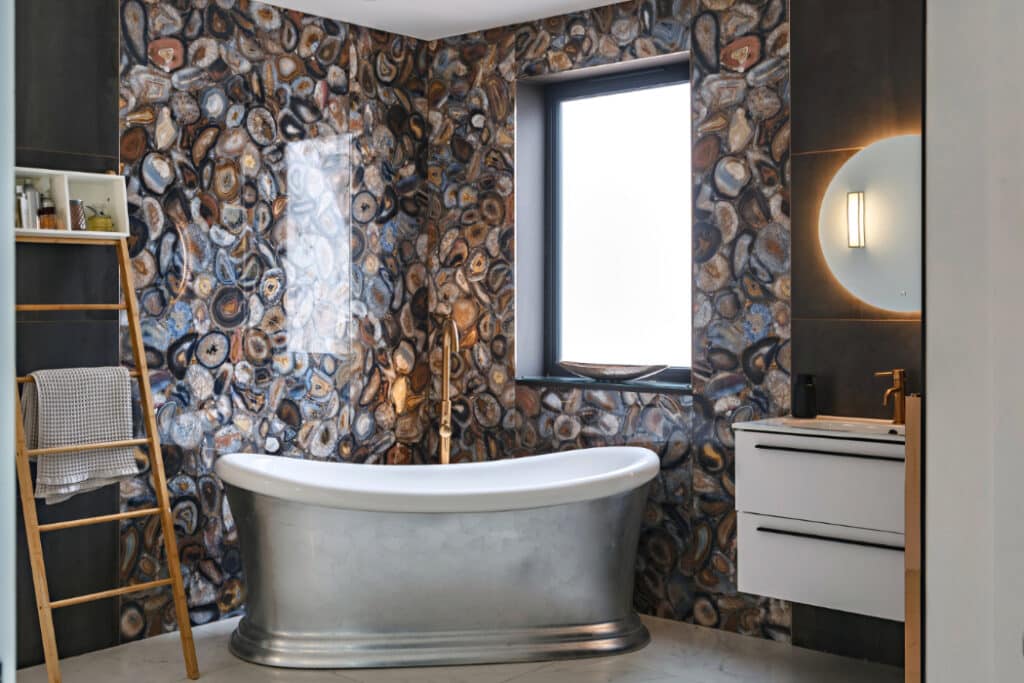 Bathroom with a freestanding silver bathtub, agate-patterned wall tiles, a ladder shelf with towels, a wall-mounted sink, and a round mirror with light above beside a window with frosted glass.