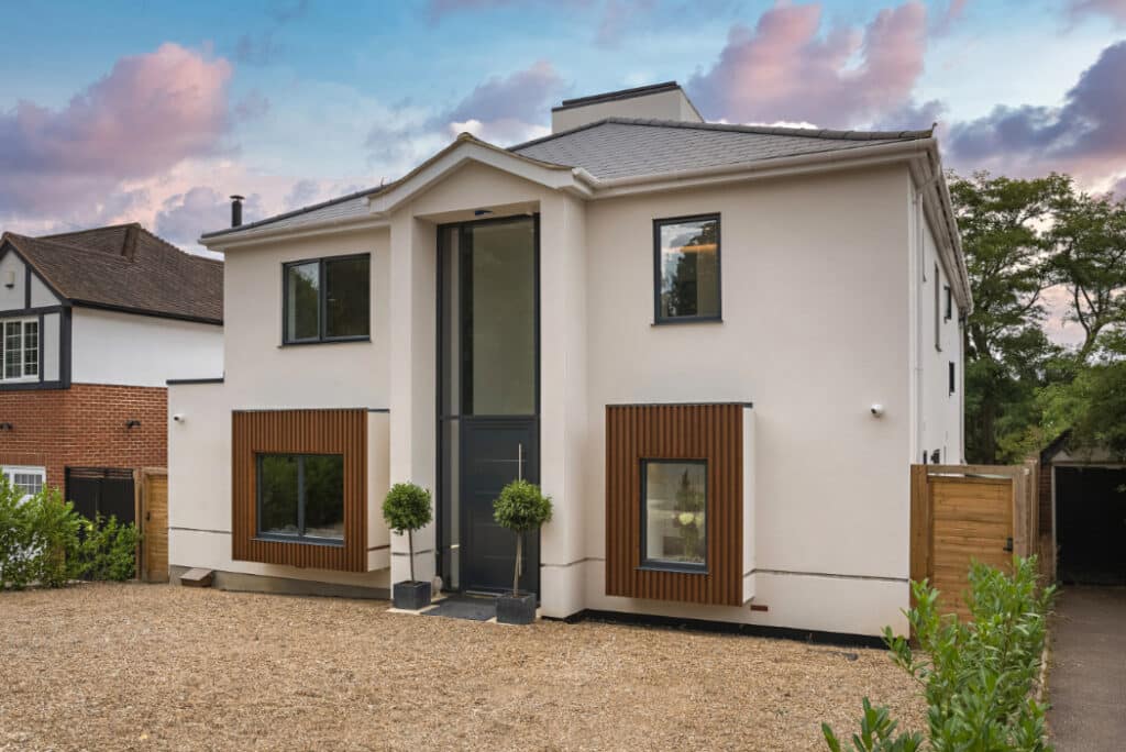 Two-story modern house with white walls, large windows, and decorative wooden panels on the first floor. A gravel driveway and small shrubs are in the foreground with a partly cloudy sky.