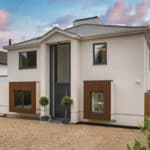 Thumbnail of http://Two-story%20modern%20house%20with%20white%20walls,%20large%20windows,%20and%20decorative%20wooden%20panels%20on%20the%20first%20floor.%20A%20gravel%20driveway%20and%20small%20shrubs%20are%20in%20the%20foreground%20with%20a%20partly%20cloudy%20sky.