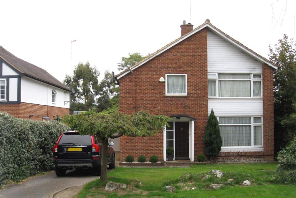 Two-story brick house with white paneling, a small front garden, and a black SUV parked in the driveway, adjacent to a green hedge.