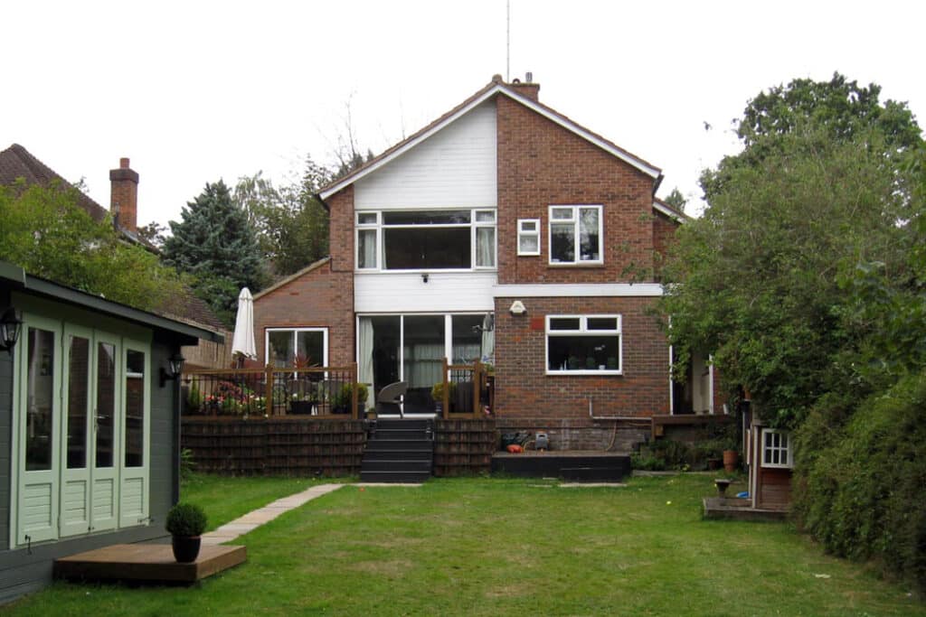 Two-story brick house with a large lawn, a raised patio with chairs on the left, and a hedge on the right. A small shed is partially visible on the right.