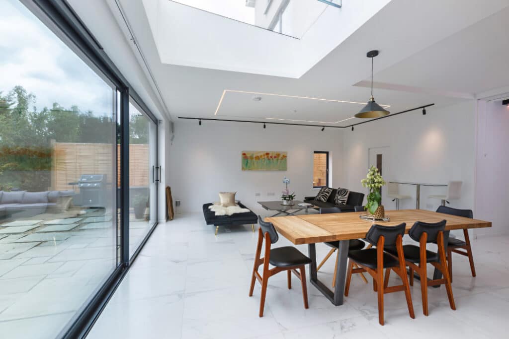 Modern dining room with a wooden table, six chairs, a black couch, and a pendant light. Large sliding glass doors open to a patio. Light decor with a painting on the wall.