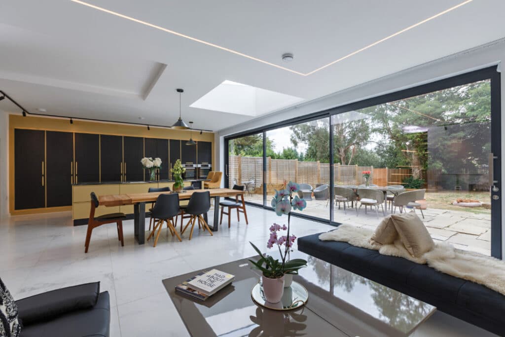 Modern open-plan kitchen and dining area with sleek furnishings, large glass sliding doors, view of a patio with outdoor seating, and a skylight.