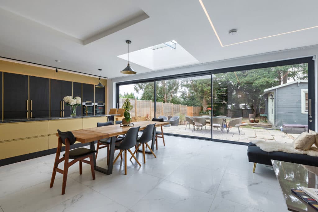 Modern kitchen and dining area with black and gold cabinets, wooden table, and sliding glass doors leading to a patio and garden.