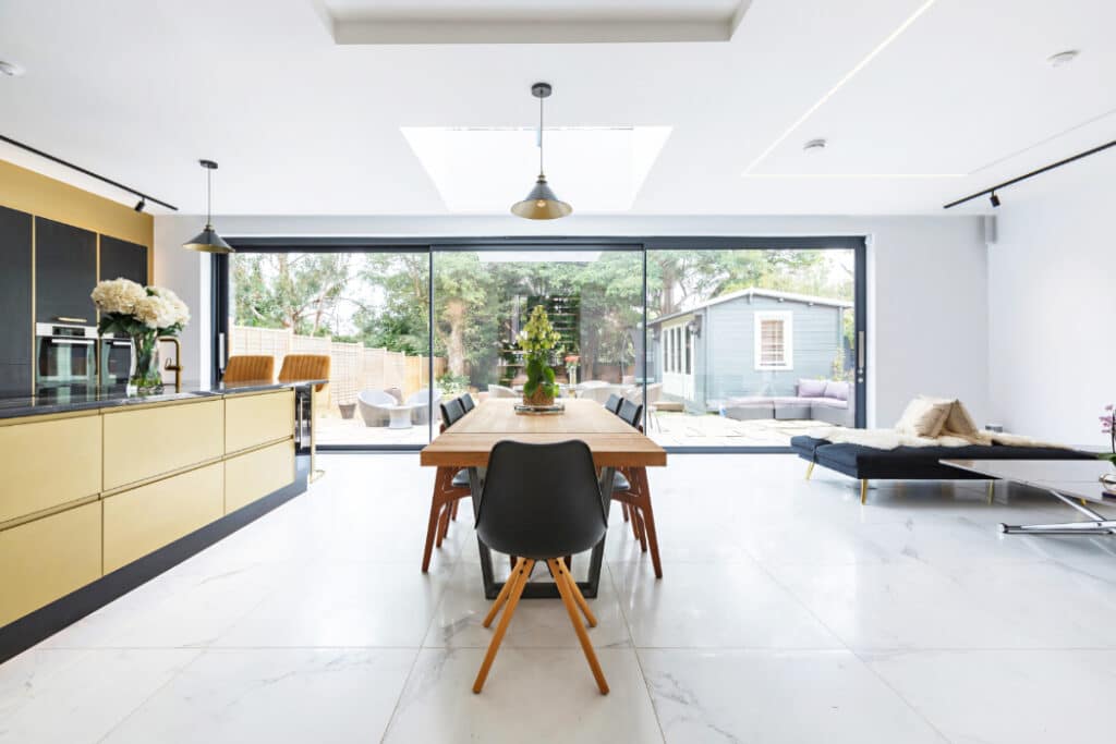 Modern kitchen and dining area with a wooden table, black chairs, gold cabinets, and large glass doors leading to a garden. Daylight illuminates the marble floor and lounge area.
