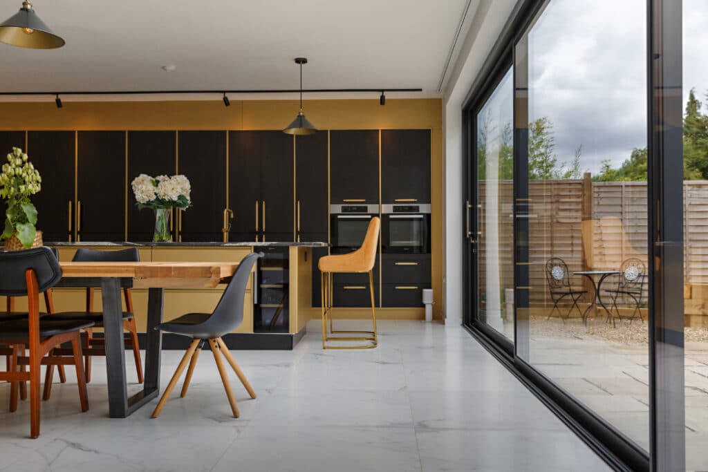 Modern kitchen with dark cabinets, a wooden dining table with mixed chairs, and large glass doors leading to an outdoor patio with a small table and two chairs. White floor tiles are visible.