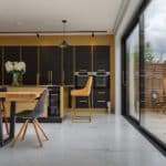 Thumbnail of http://Modern%20kitchen%20with%20dark%20cabinets,%20a%20wooden%20dining%20table%20with%20mixed%20chairs,%20and%20large%20glass%20doors%20leading%20to%20an%20outdoor%20patio%20with%20a%20small%20table%20and%20two%20chairs.%20White%20floor%20tiles%20are%20visible.