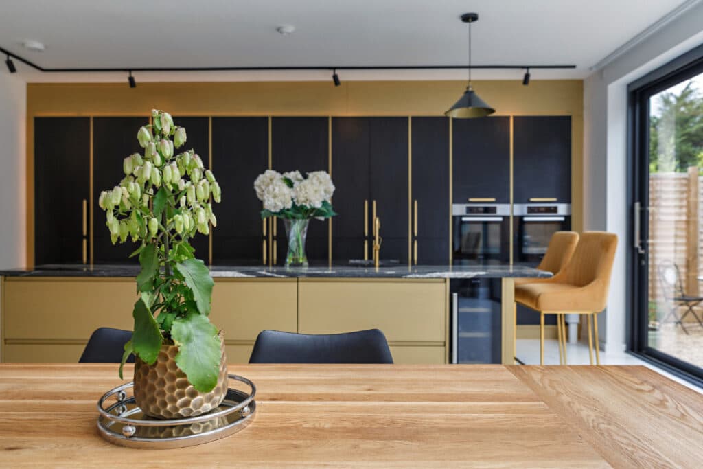 Modern kitchen with wooden dining table, potted plant centerpiece, gold and black cabinetry, double oven, and two mustard yellow chairs near large windows.