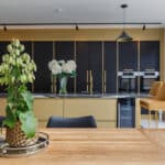 Thumbnail of http://Modern%20kitchen%20with%20wooden%20dining%20table,%20potted%20plant%20centerpiece,%20gold%20and%20black%20cabinetry,%20double%20oven,%20and%20two%20mustard%20yellow%20chairs%20near%20large%20windows.