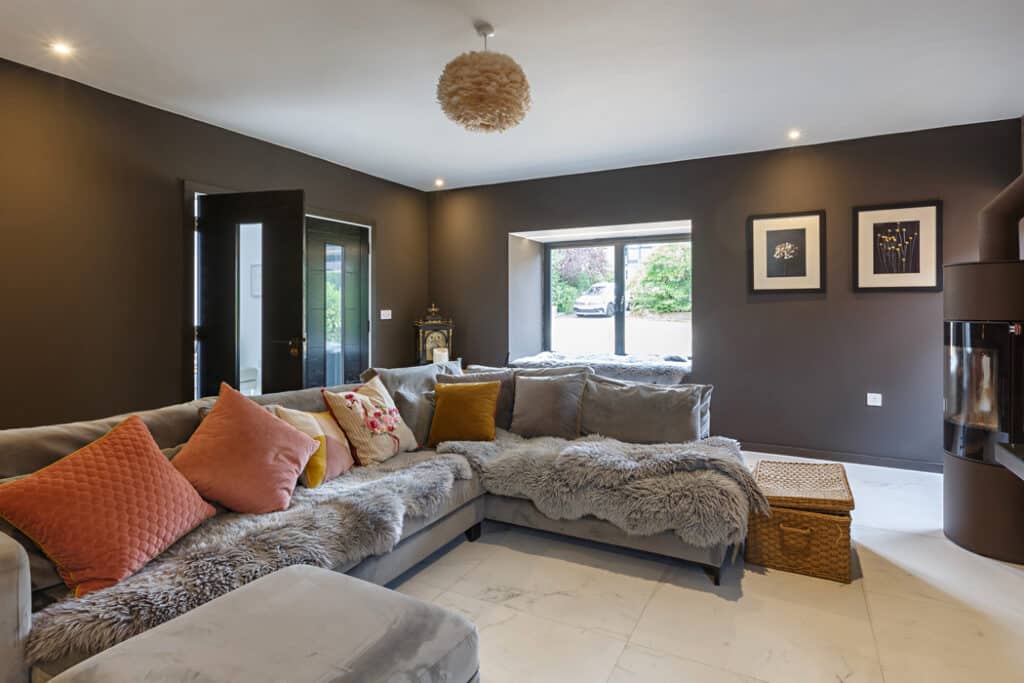 Modern living room with a gray sectional sofa adorned with colorful pillows and a fur throw. A wicker basket serves as a table. The walls are dark with framed art and a window to the outside.