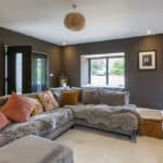 Thumbnail of http://Modern%20living%20room%20with%20a%20gray%20sectional%20sofa%20adorned%20with%20colorful%20pillows%20and%20a%20fur%20throw.%20A%20wicker%20basket%20serves%20as%20a%20table.%20The%20walls%20are%20dark%20with%20framed%20art%20and%20a%20window%20to%20the%20outside.