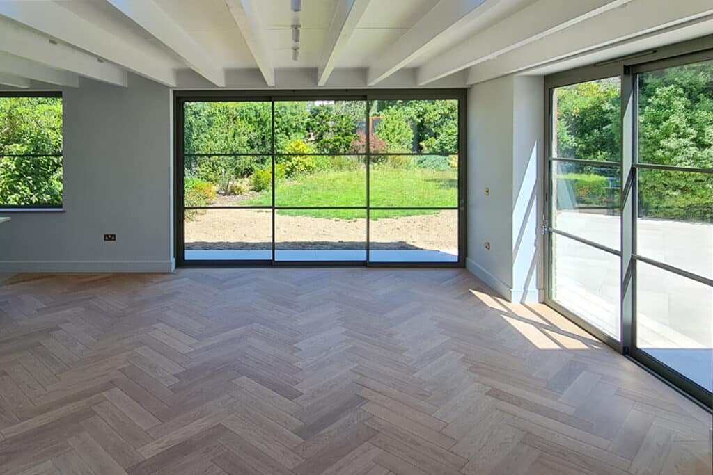 Bright, empty room with large floor-to-ceiling windows overlooking a garden. Light wood herringbone floor, white walls, and exposed ceiling beams are visible.