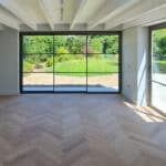 Thumbnail of http://Bright,%20empty%20room%20with%20large%20floor-to-ceiling%20windows%20overlooking%20a%20garden.%20Light%20wood%20herringbone%20floor,%20white%20walls,%20and%20exposed%20ceiling%20beams%20are%20visible.