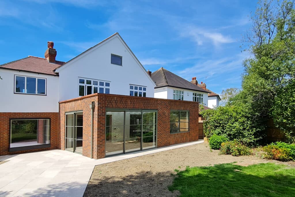 A modern house with red brick and white facade, large glass doors, and a small garden under a clear blue sky.