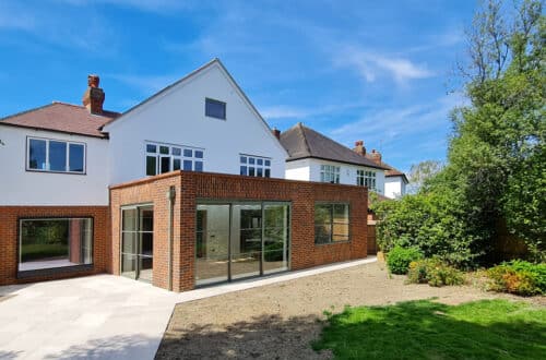 A modern house with red brick and white facade, large glass doors, and a small garden under a clear blue sky.