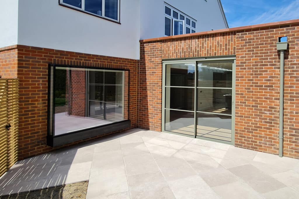 A modern brick house with large glass sliding doors and windows, leading to a spacious tiled patio under a clear blue sky.