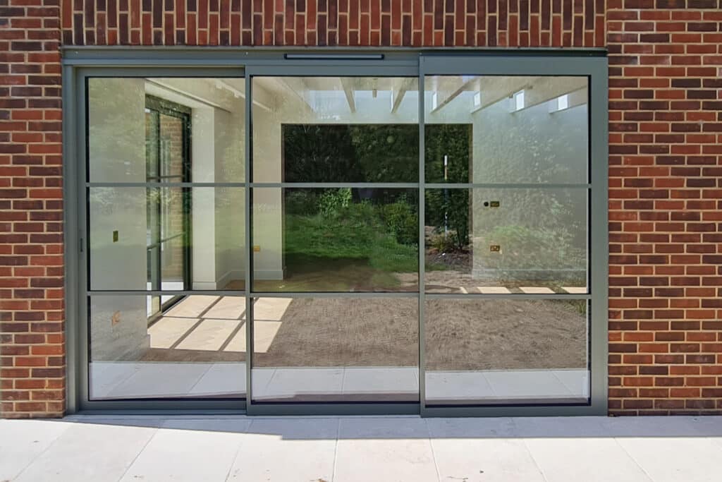 Sliding glass patio doors with grid-pattern frames on a brick wall, reflecting a green outdoor area.