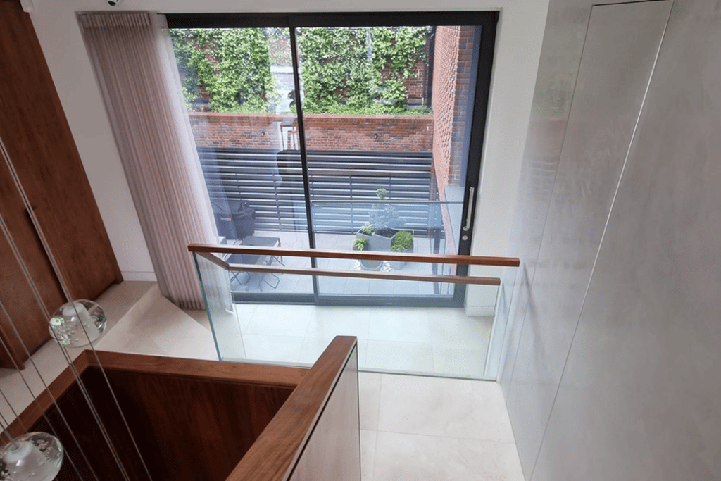 View from a staircase landing showing a glass door leading to a patio with potted plants and a small table with chairs. A brick wall with greenery is visible outside.