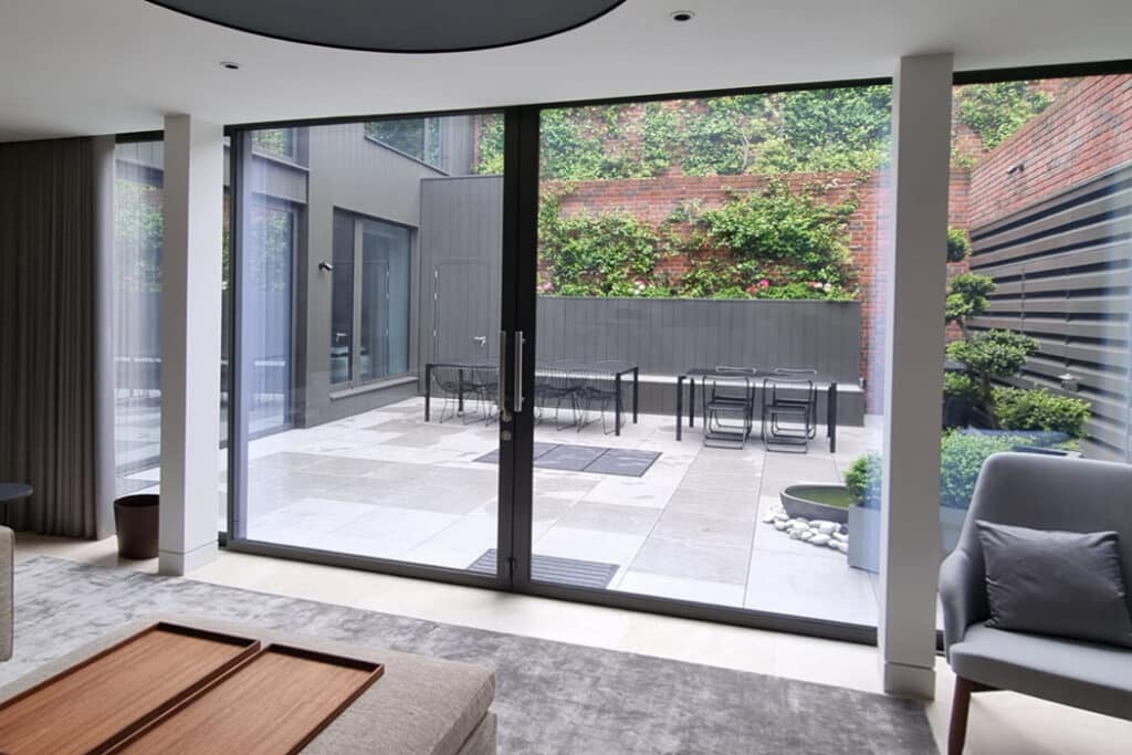 Modern living room with large glass doors opening to a patio. The patio features outdoor furniture, plants, and vertical wooden slats against a brick wall.