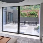 Thumbnail of http://Modern%20living%20room%20with%20large%20glass%20doors%20opening%20to%20a%20patio.%20The%20patio%20features%20outdoor%20furniture,%20plants,%20and%20vertical%20wooden%20slats%20against%20a%20brick%20wall.