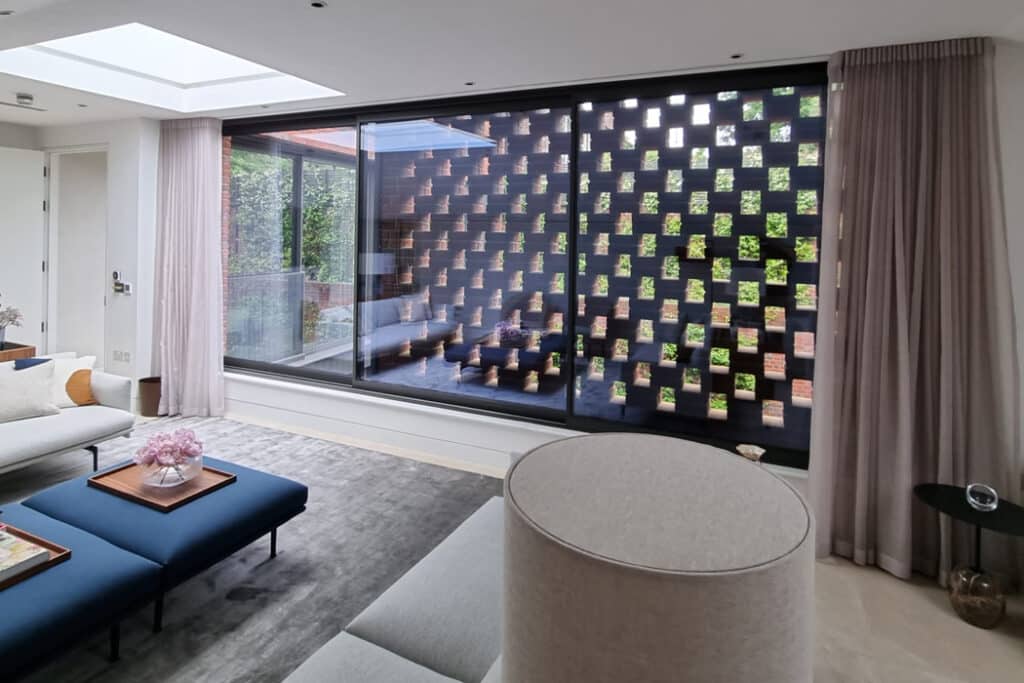 Modern living room with large window featuring a grid-patterned screen. White walls, gray rug, blue ottomans, and a round beige chair are visible. Pink flowers adorn a table.