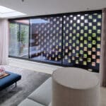 Thumbnail of http://Modern%20living%20room%20with%20large%20window%20featuring%20a%20grid-patterned%20screen.%20White%20walls,%20gray%20rug,%20blue%20ottomans,%20and%20a%20round%20beige%20chair%20are%20visible.%20Pink%20flowers%20adorn%20a%20table.