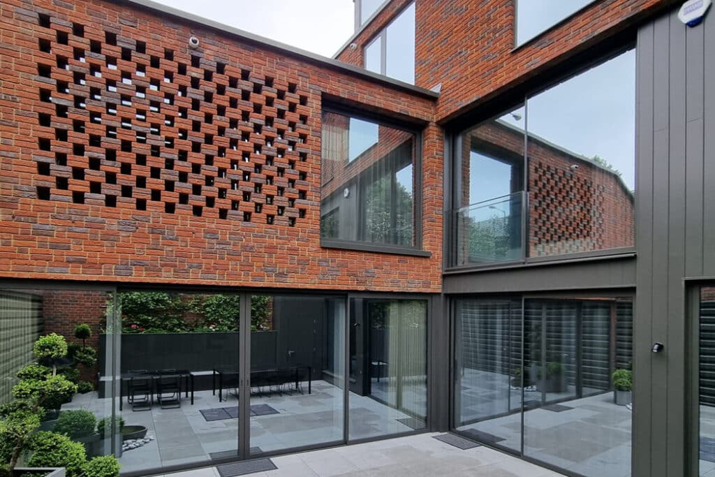 Modern architectural exterior featuring brick latticework, large glass windows, and a courtyard with potted plants and outdoor seating.