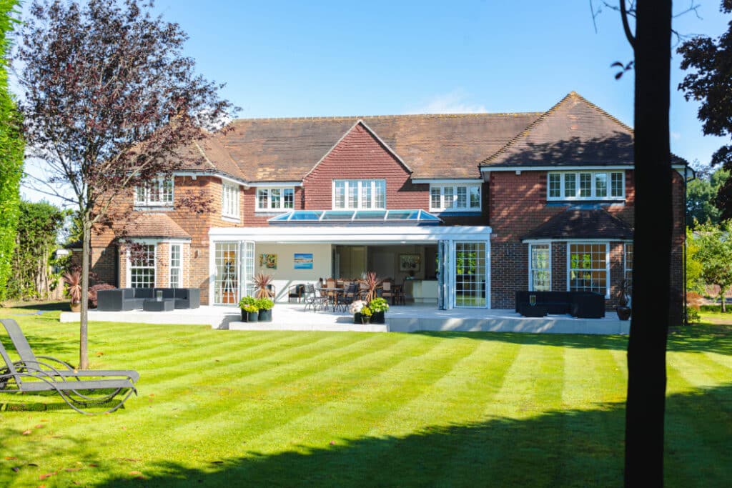 A large brick house with a sunroom and patio, surrounded by a neatly manicured lawn and trees on a clear, sunny day.