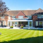 Thumbnail of http://A%20large%20brick%20house%20with%20a%20sunroom%20and%20patio,%20surrounded%20by%20a%20neatly%20manicured%20lawn%20and%20trees%20on%20a%20clear,%20sunny%20day.