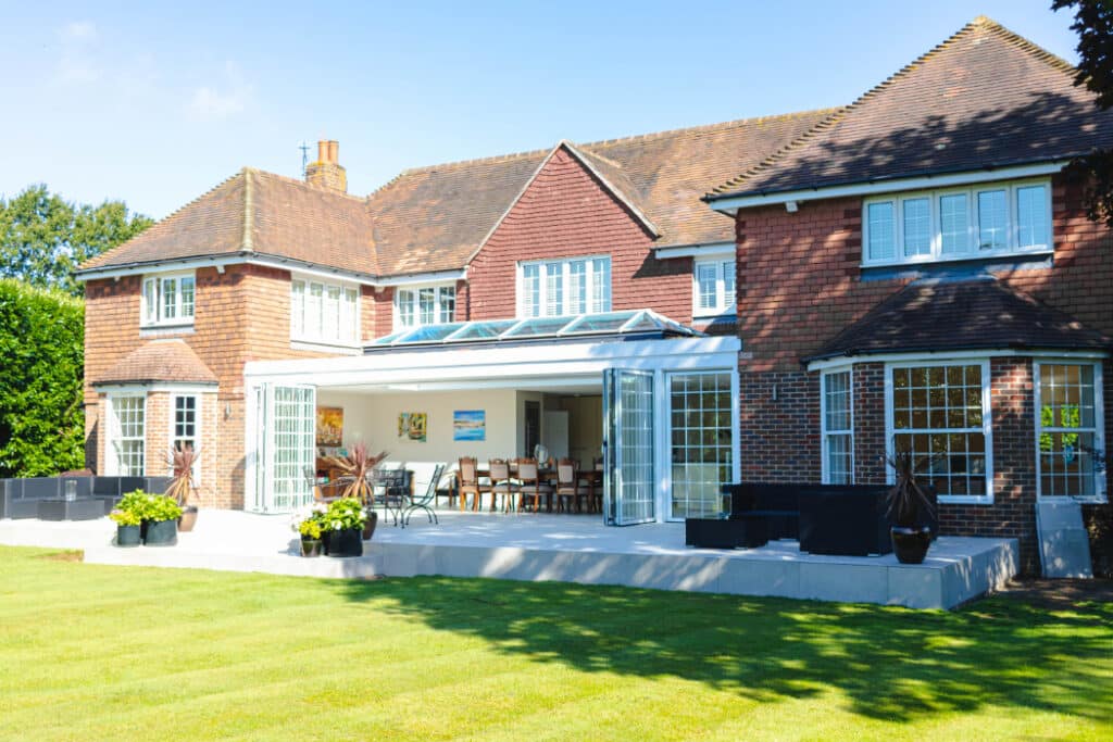 Large brick house with an open sunroom, expansive glass doors, and a spacious patio overlooking a manicured lawn.