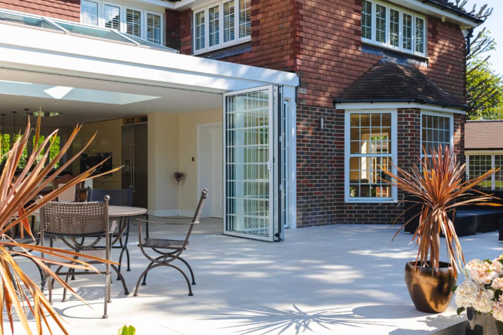 A modern house exterior with large folding glass doors, leading to a spacious patio. Outdoor furniture and potted plants are arranged on the patio.