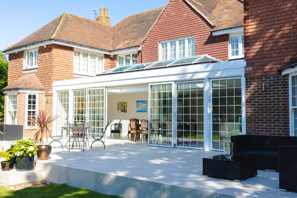 A large brick house with a glass conservatory and sliding doors opens onto a spacious patio with outdoor furniture.