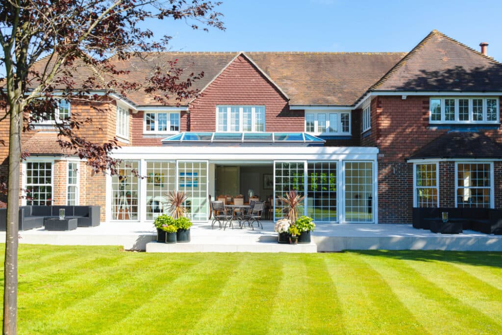 A large brick house with a glass sunroom extension, surrounded by a well-maintained lawn and plant pots.