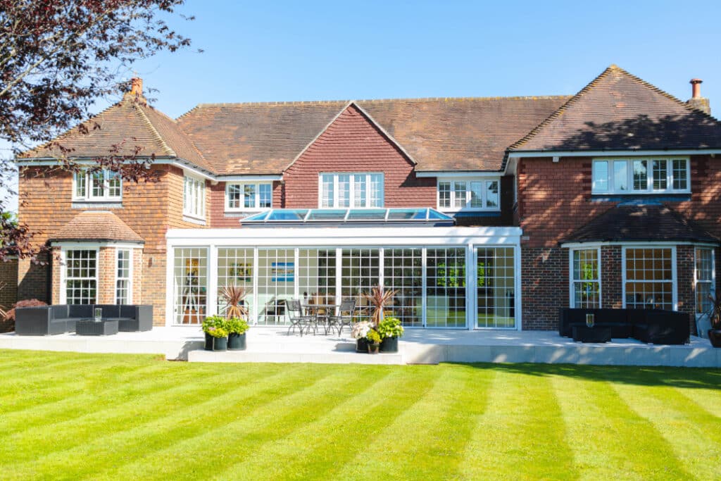 A large brick house with multiple sections, featuring a glass conservatory and several windows, set against a well-maintained lawn and clear blue sky.