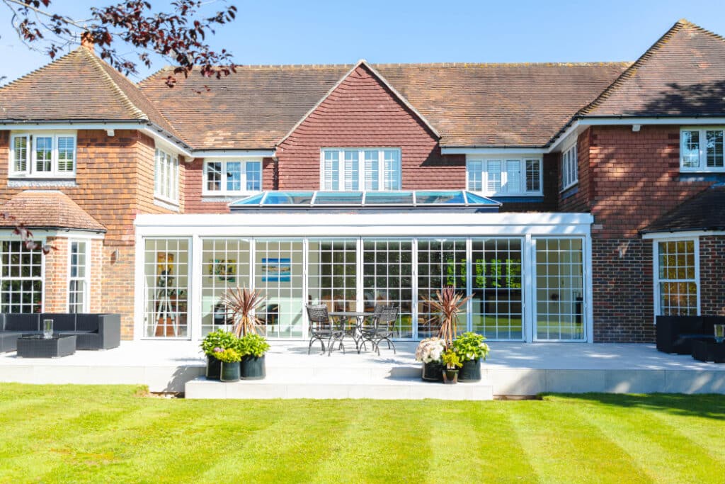 Large brick house with a glass-enclosed patio, surrounded by a well-manicured lawn. Outdoor furniture is visible on the patio.