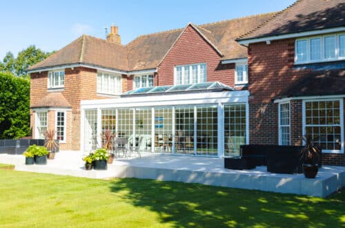 Brick house with a glass conservatory extending into a patio, surrounded by a lawn with potted plants.