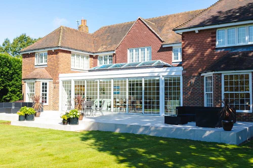 Brick house with a glass conservatory extending into a patio, surrounded by a lawn with potted plants.